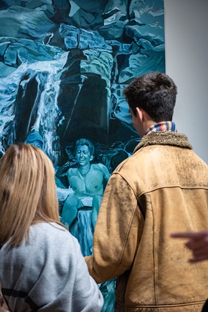 Two people stand in front of a blue-tinged painting of a shirtless young man standing underneath a waterfall.