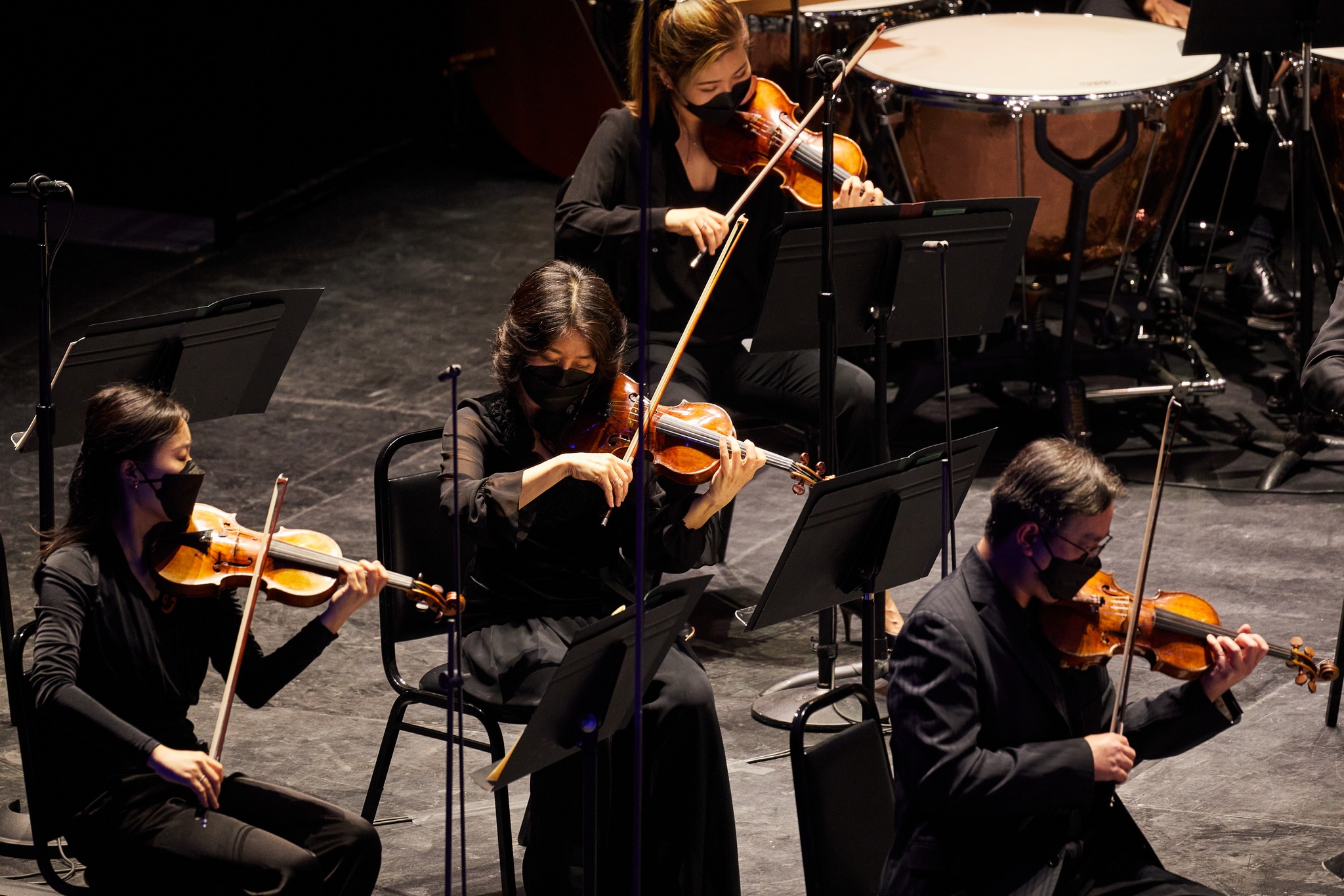 Four violinists on a black stage playing their instruments while wearing black clothes and matching face masks