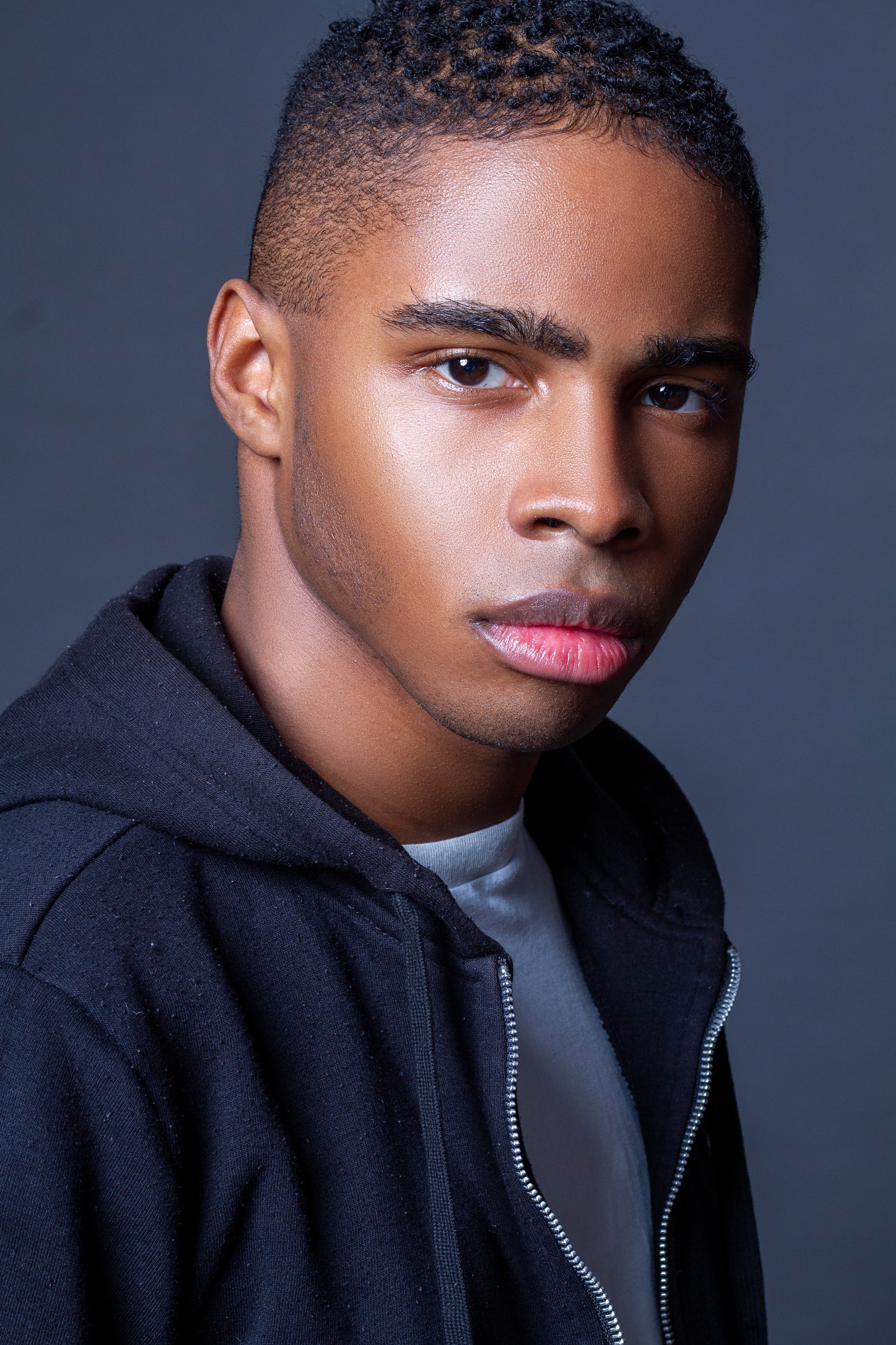 A Black man seen in profile turns his face toward the camera. He has a serious expression and wears a zip-up hoodie over a blue t-shirt. 