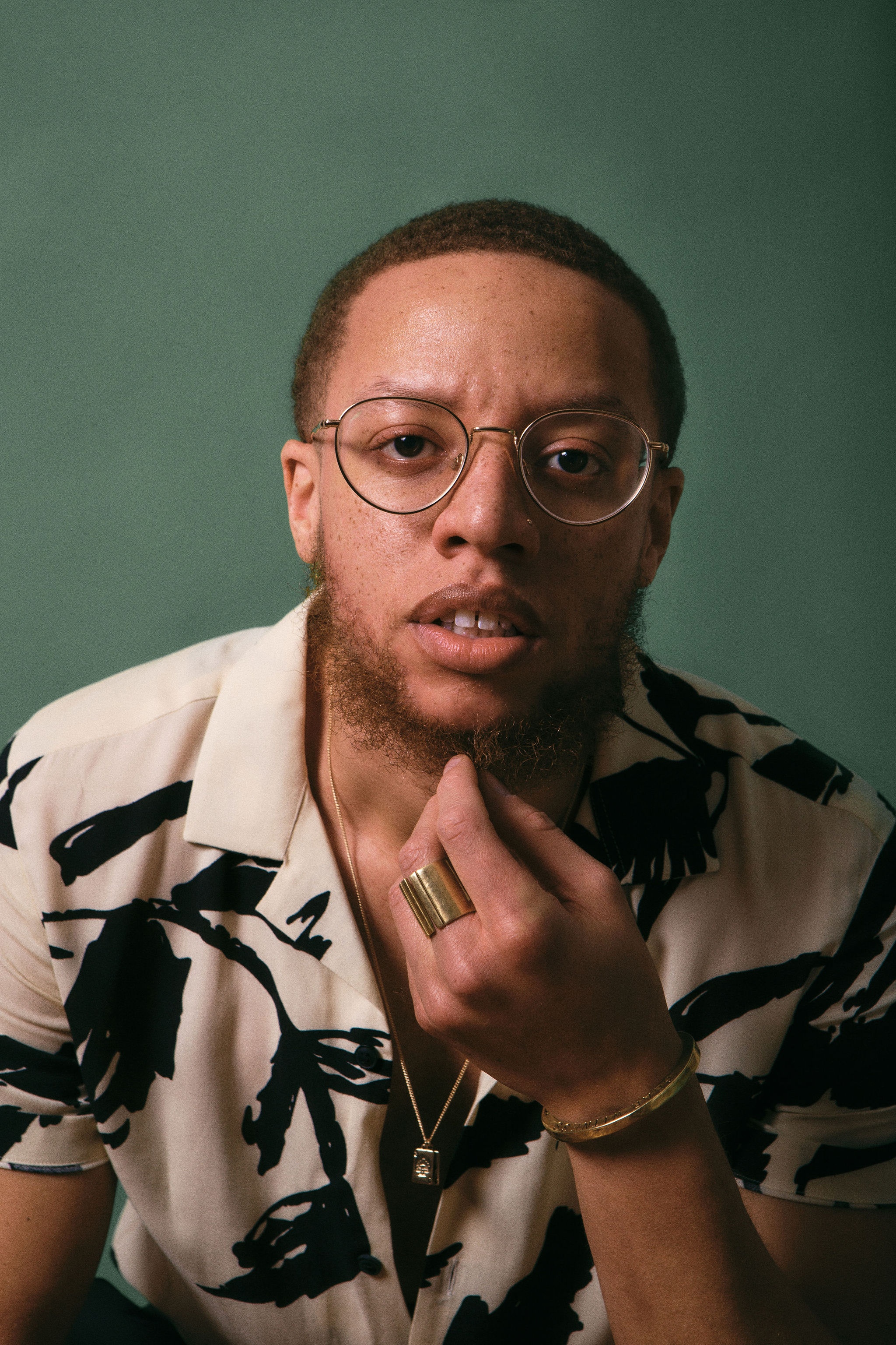 A portrait of Najee Omar, a queer Black person who poses in front of a khaki green backdrop. He leans forward with one hand to his chin. He has light brown skin, a slightly bushy light brown beard and wears glasses. Photo by Christian Hicks