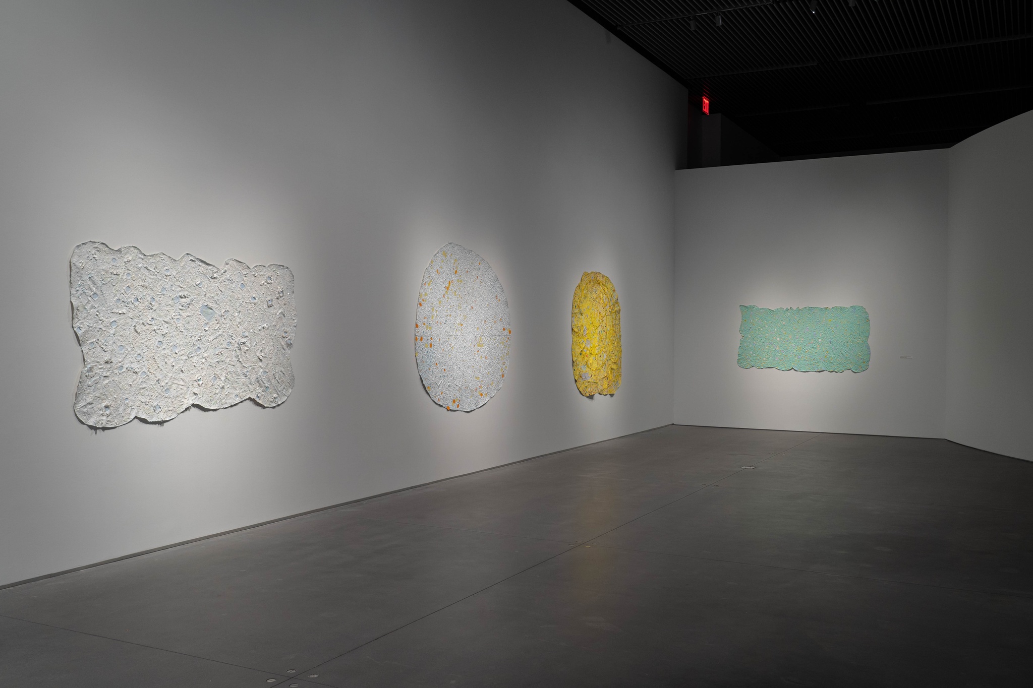 An installation view of abstract paintings in the exhibition Howardena Pindell: Rope/Fire/Water. The curvilinear and rectangular canvases progress from two works predominantly in white, to a yellow canvas, to a green rectangular painting with irregular edges facing the camera on the far wall.