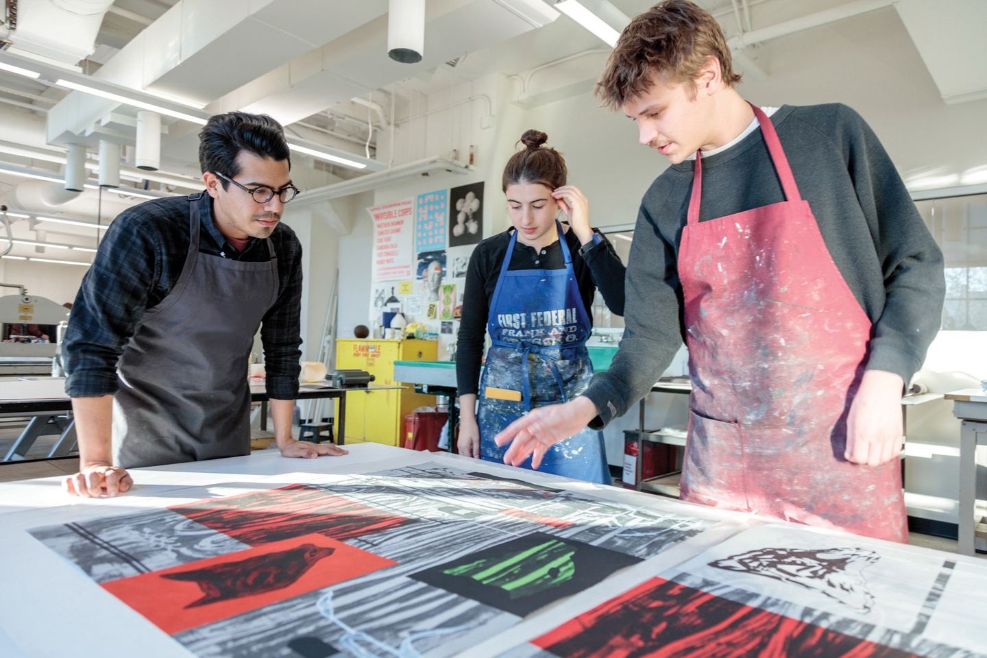 Three students in aprons standing in front of a collection of colorful prints in a spacious printmaking studio.