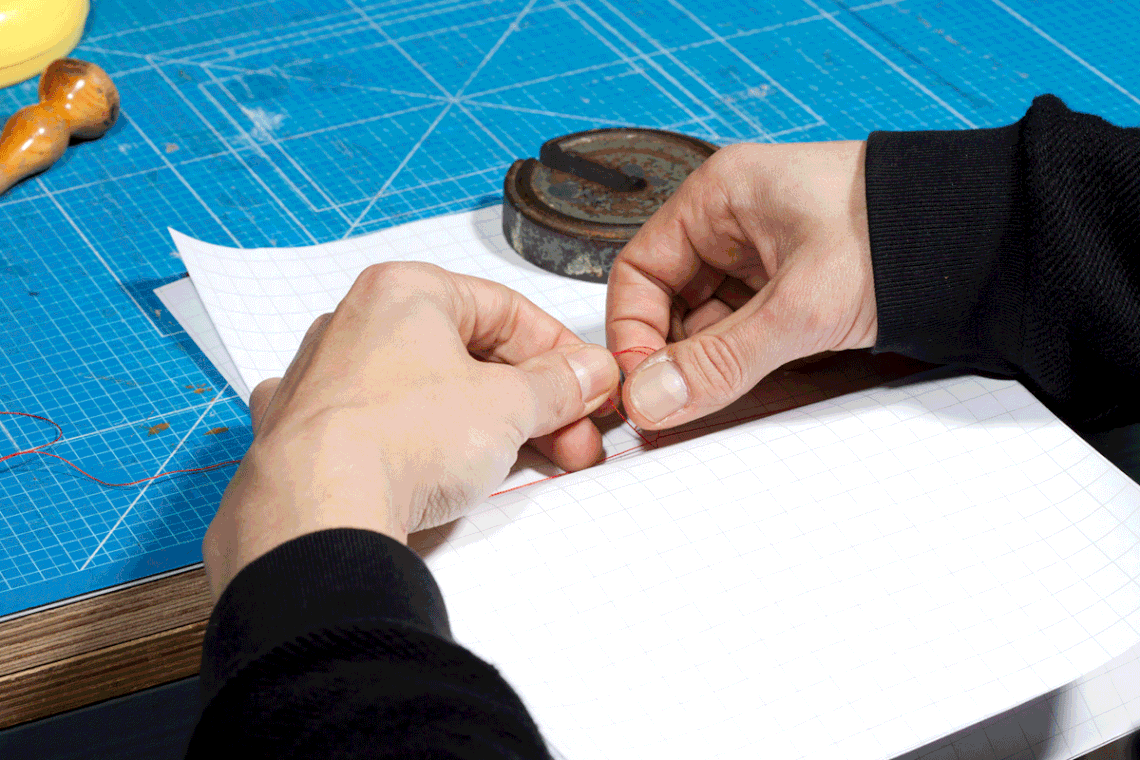 .GIF of light-skinned hands tying a knot with red twine on the binding of a blank book on a bright blue linoleum mat.