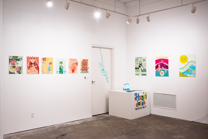 Wide shot of gallery space with nine illustrated poster designs, a display stand with a fabricated resort sign, a stack of brochures, and a vinyl decal of a person swinging on a rope on the wall.