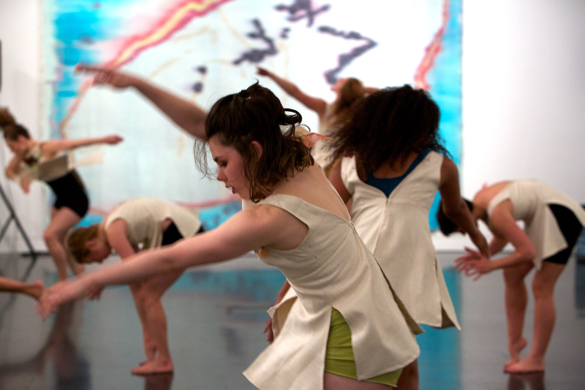 A white, female dancer caught in motion in front of a group of dancers in front of a large blue, red, white, yellow, and purple painting.