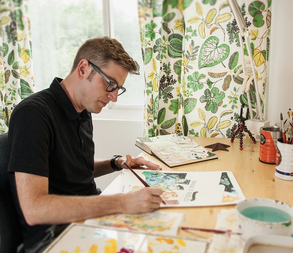 John Hendrix seated at a desk, working on illustration work in front of him, brush in hand. 