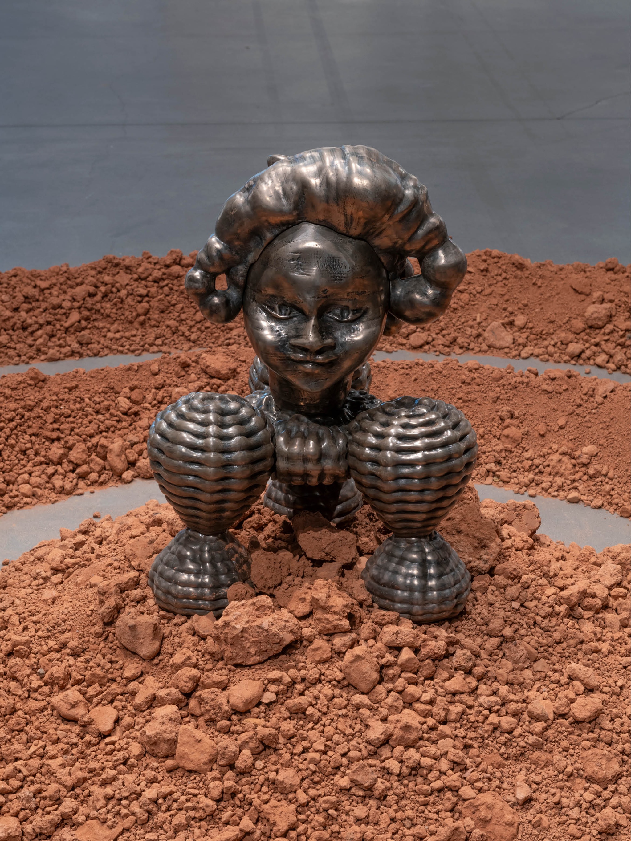 A wide sculptural artwork on a gallery floor. It consists of two concentric rings of red dirt. At the center on top of a mound of the dirt sits a bronze sculpture inspired by the Benin Bronzes, historical artworks from Benin, present-day Nigeria.
