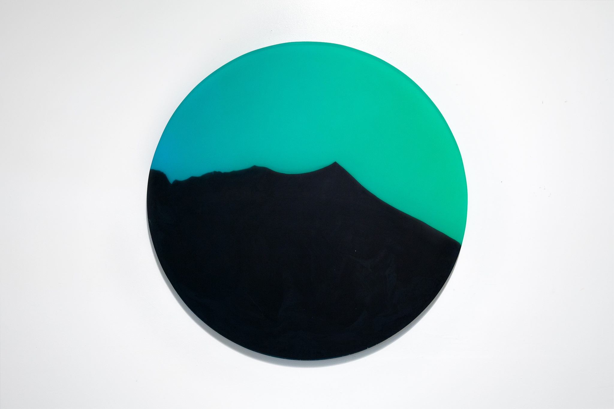 A disk sits on a white background. The top half of the disk is a mix of blue-green colors and the bottom half is defined by a black section with a jagged point that pushes into the top half of the disk, as if depicting a mountain landscape in shadow against a blue-green sky.
