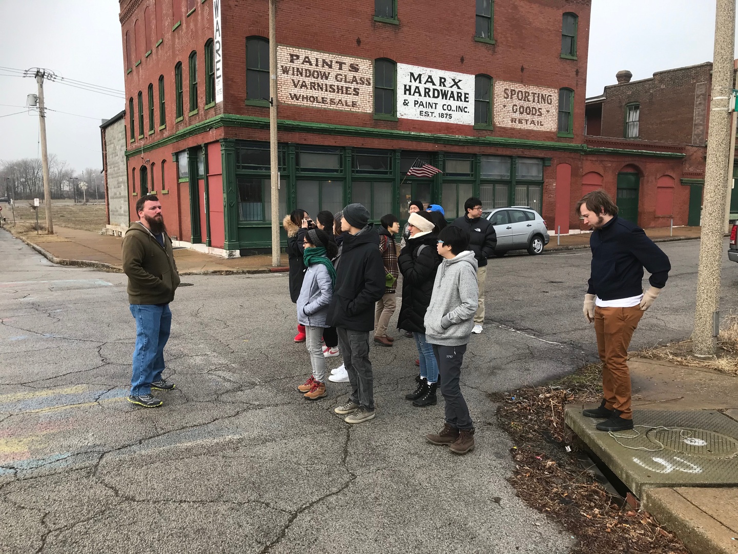 A group of people stand on the street and sidewalk in front of a brick building with the sign "Marx Hardware"