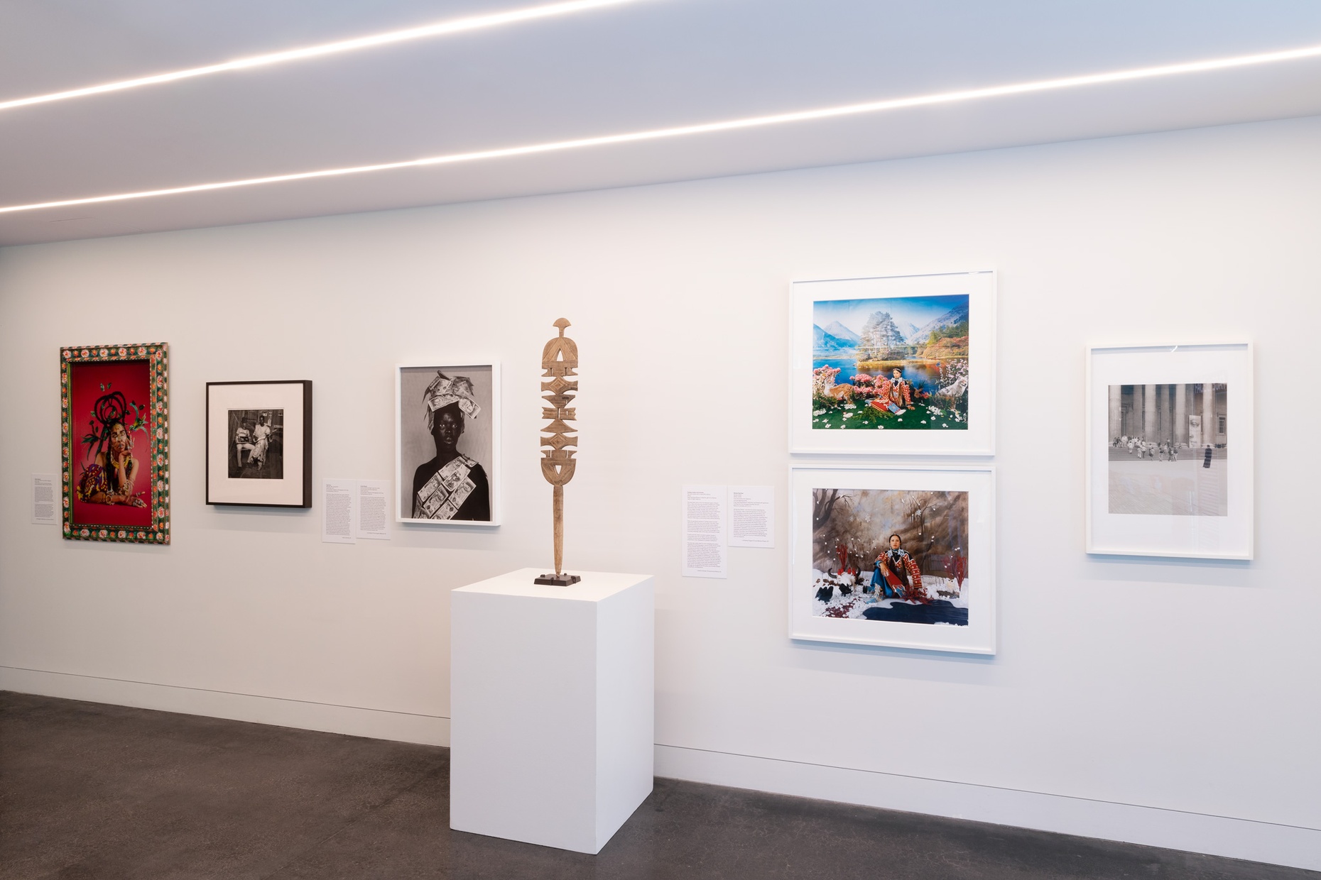 A gallery wall displaying multiple framed photographs and one wooden sculpture on a pedestal.