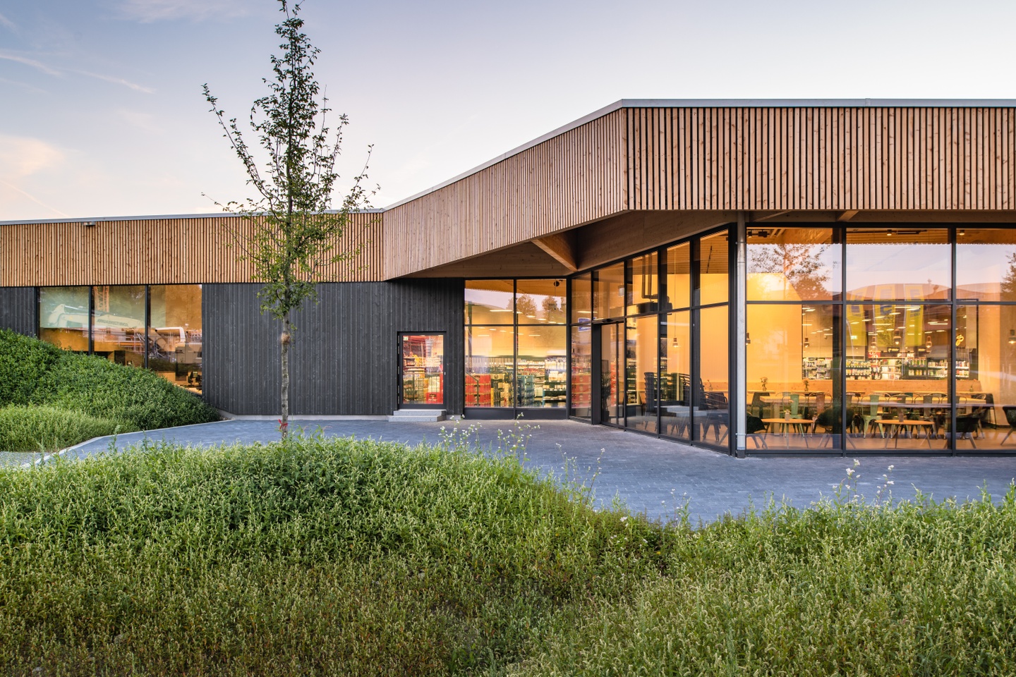 Exterior of a modern supermarket surrounded by prairie grass and a walking path. The building has floor the ceiling windows and wooden accents. Inside can be seen shelves and a cafeteria dining area.