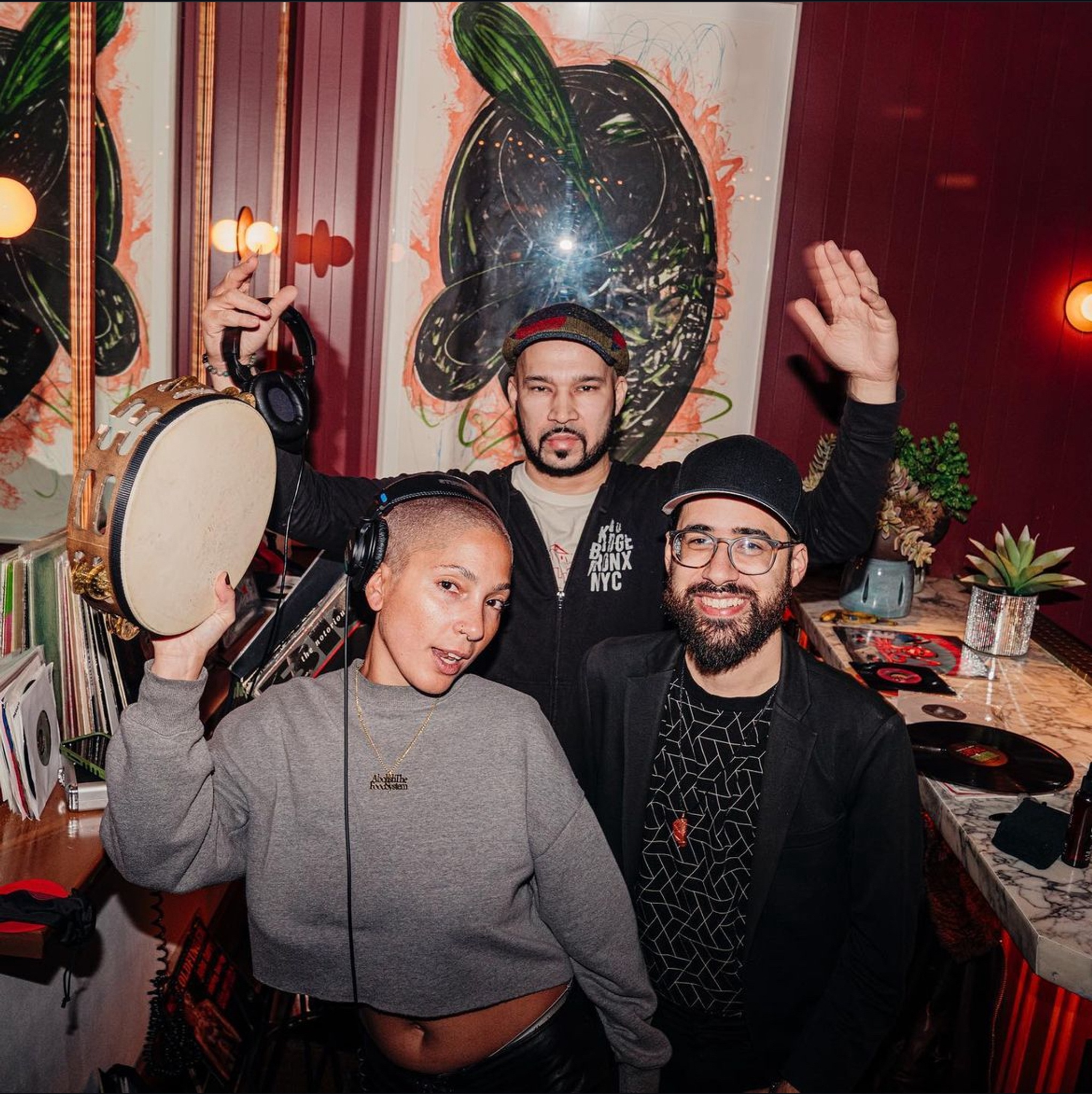 The three DJs of Uptown Vinyl Supreme, Sunny Cheeba, Buddy, and Josh Hubi, stand together in a DJ booth. They smile and raise their hands, holding headphones and tambourines. 