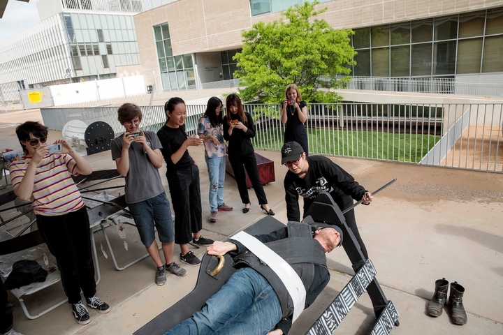 6 students observe and take photos on their phones of a student rotating a secured-by-straps Arny like a rotisserie chicken