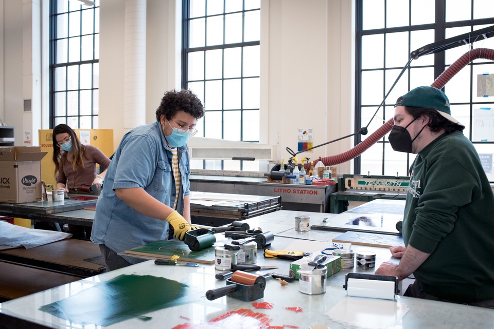 Two people chatting while one applies ink to a relief printing block with a brayer.