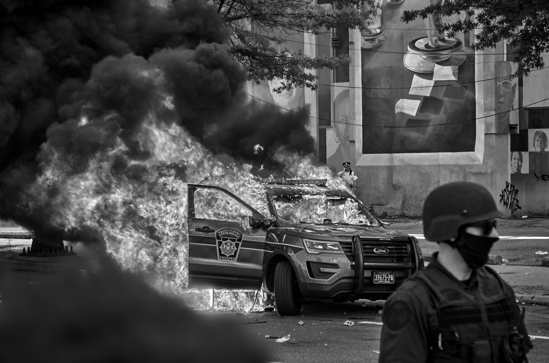 A black and white photograph of a police car on fire in front of a wall with a mural.