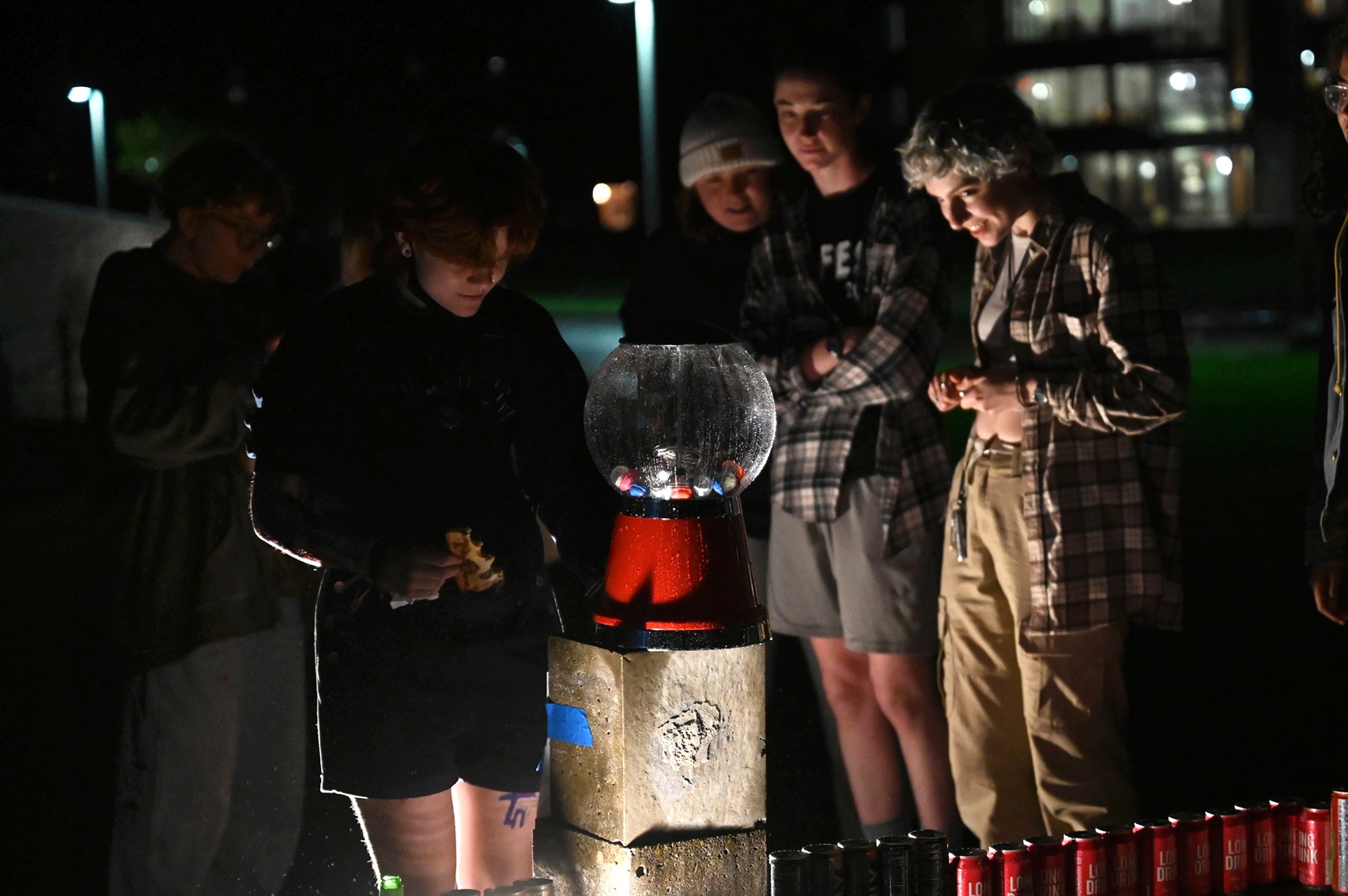 Outside at night, people gather around a gum ball machine.