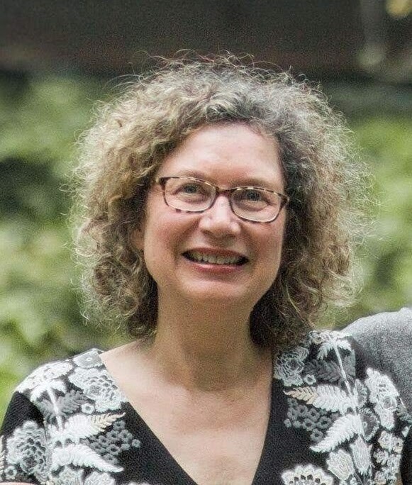A woman with curly hair and glasses smiles against an outside background of blurry green leaves. 