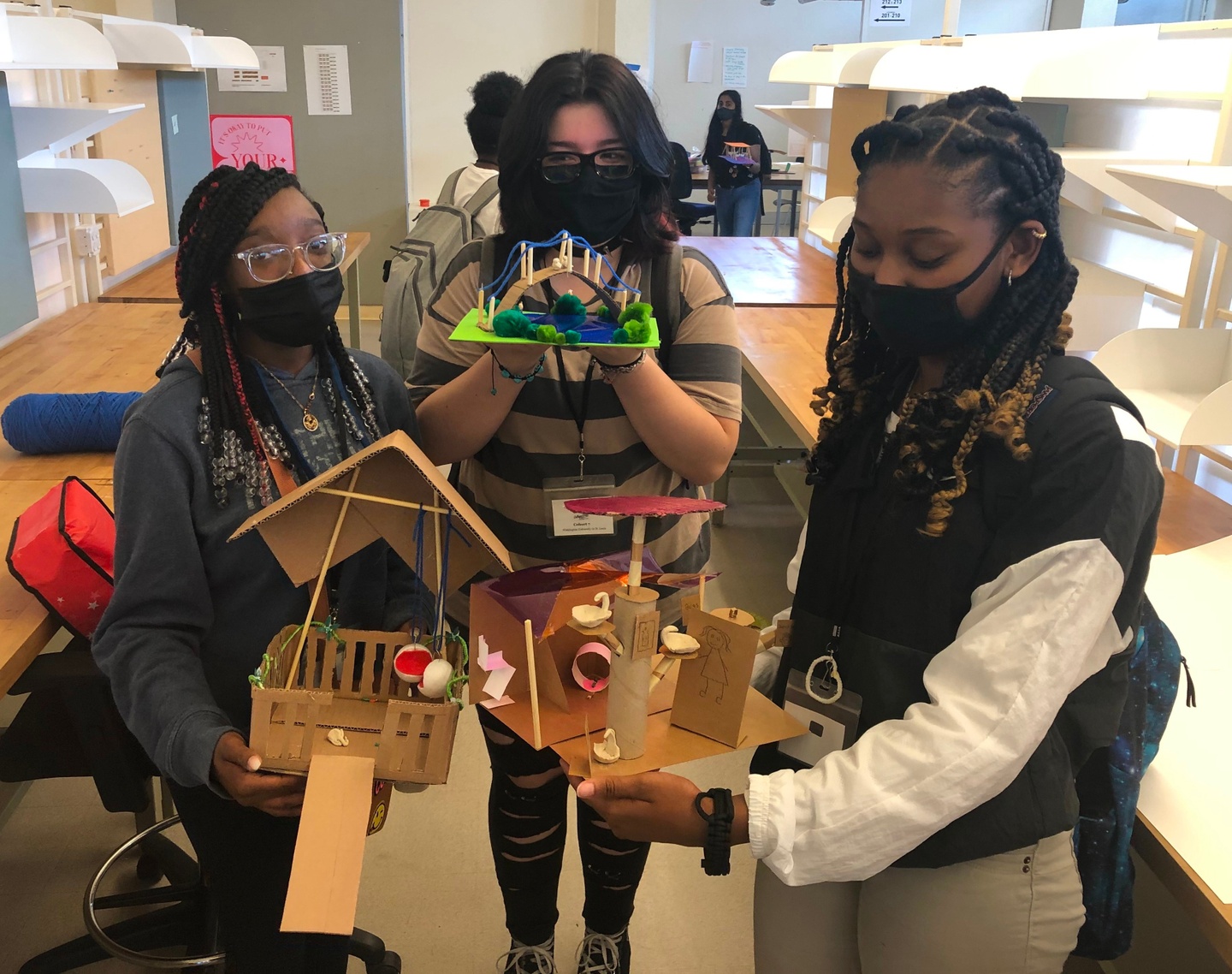 A group of three people wearing masks stands between desks, holding models made of wood and paper. 