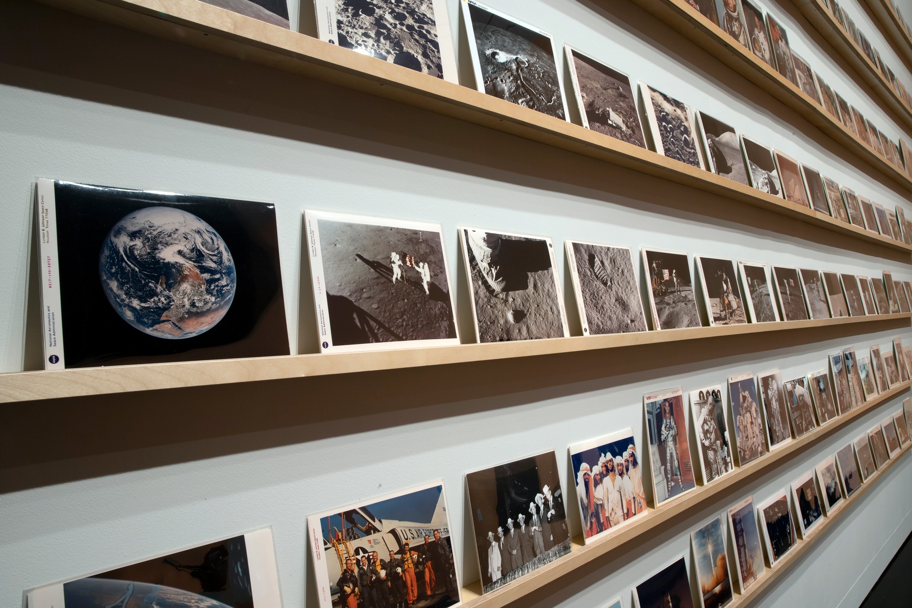 Seen at an angle, a white wall features multiple rows of many small photographs sitting on long, narrow wooden shelves.