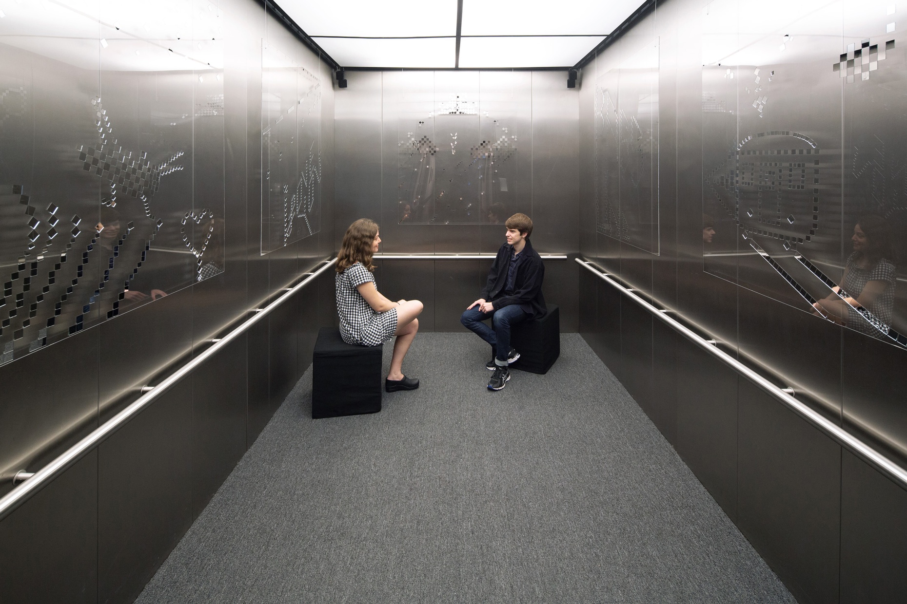 Two people sit on stools in an elevator space with art made of plexiglass and mirrors hanging on the walls.