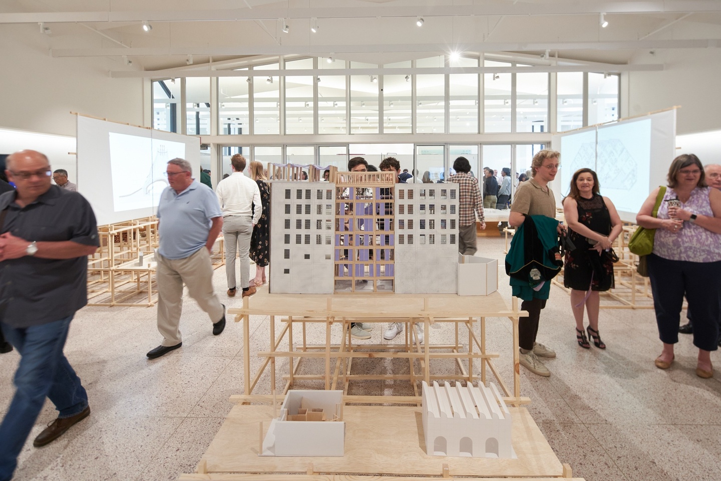 Gallery space with a row of architectural models displayed along the center aisle, and two freestanding banks of projector screens flaking the sides, displaying architectural renderings. Attendees walk through the space.
