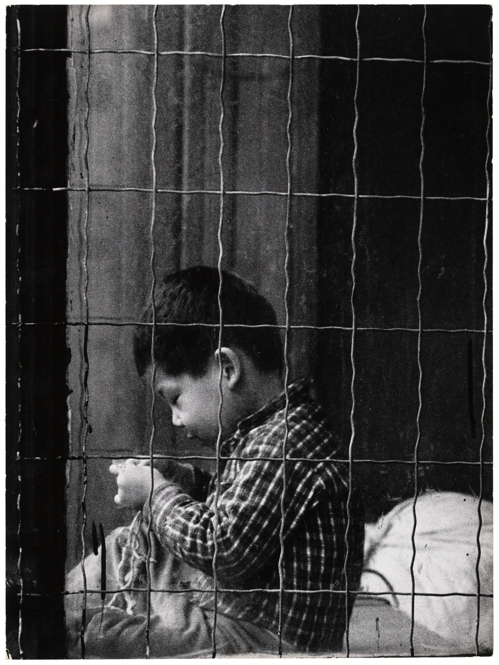 A black and white photograph taken through a gridded security window of a small child in profile working with string in his hand.