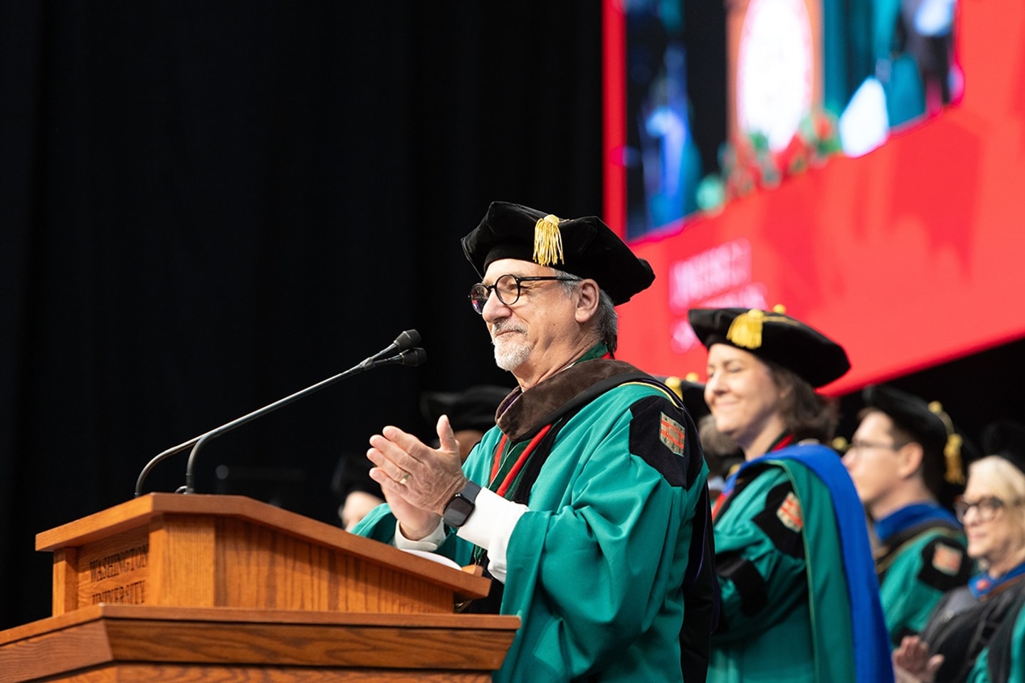 A man with glasses wearing regalia stands at a lectern and claps while looking at the audience.