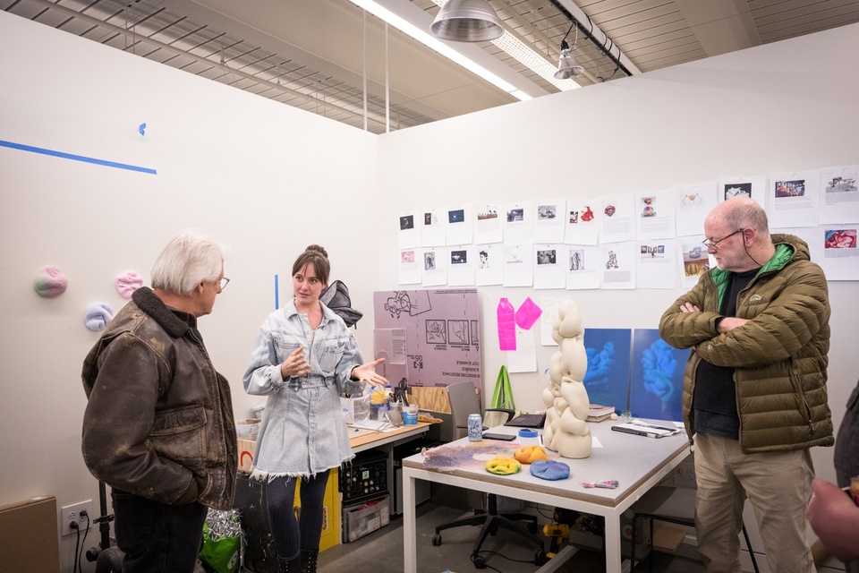 Student explains something to two visitors in a studio space. On the table are some smooth, rounded sculptural objects and pinned to the wall are a series of reference images.