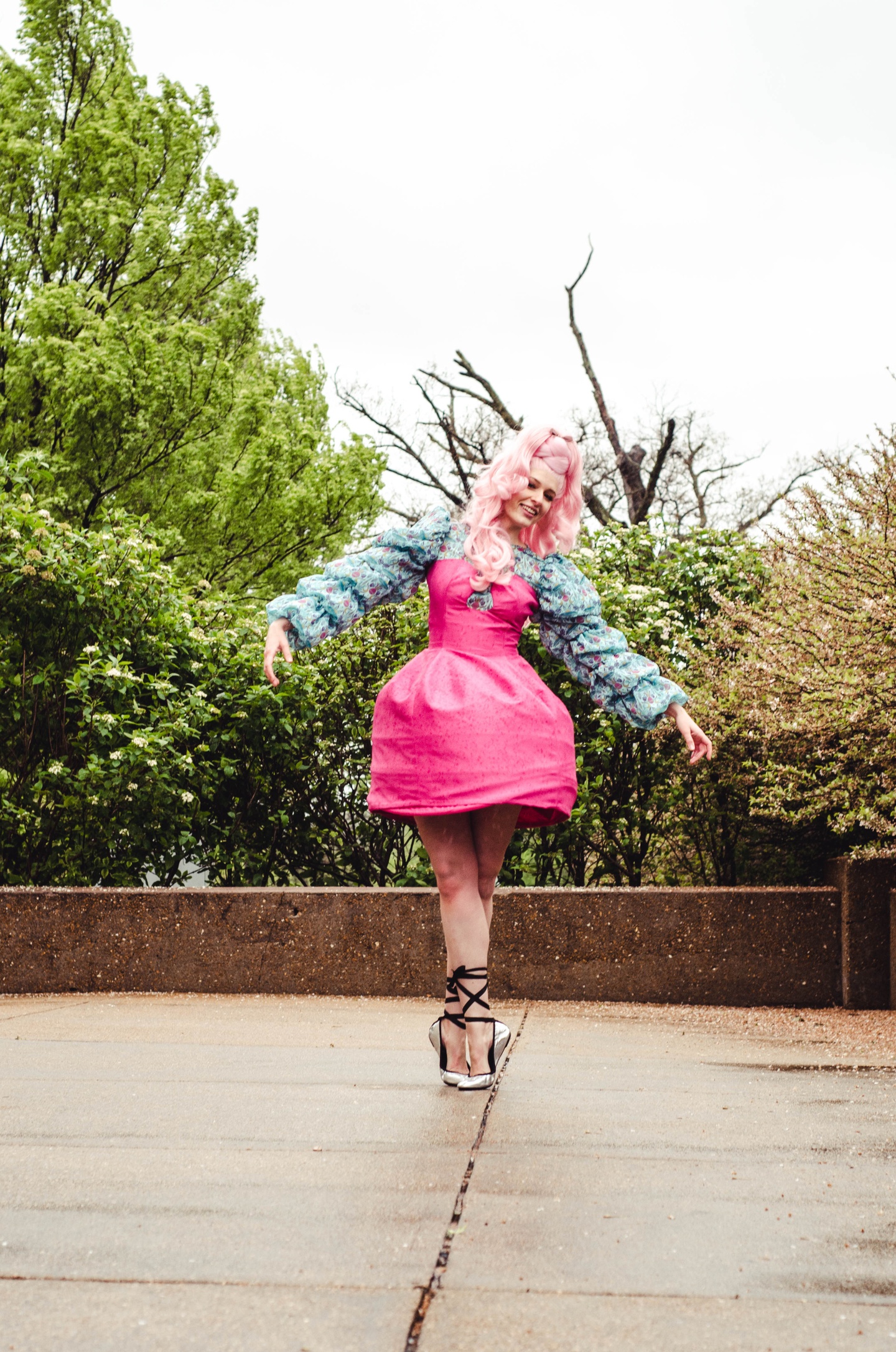 Model in a pink curly wig wears a hot pink thigh-length dress with a stiff, balloon-shaped skirt and a corseted top, over a long sleeve ruffled shirt made of blue patterned fabric. The model wears silver ballet shoes and executes pointe.