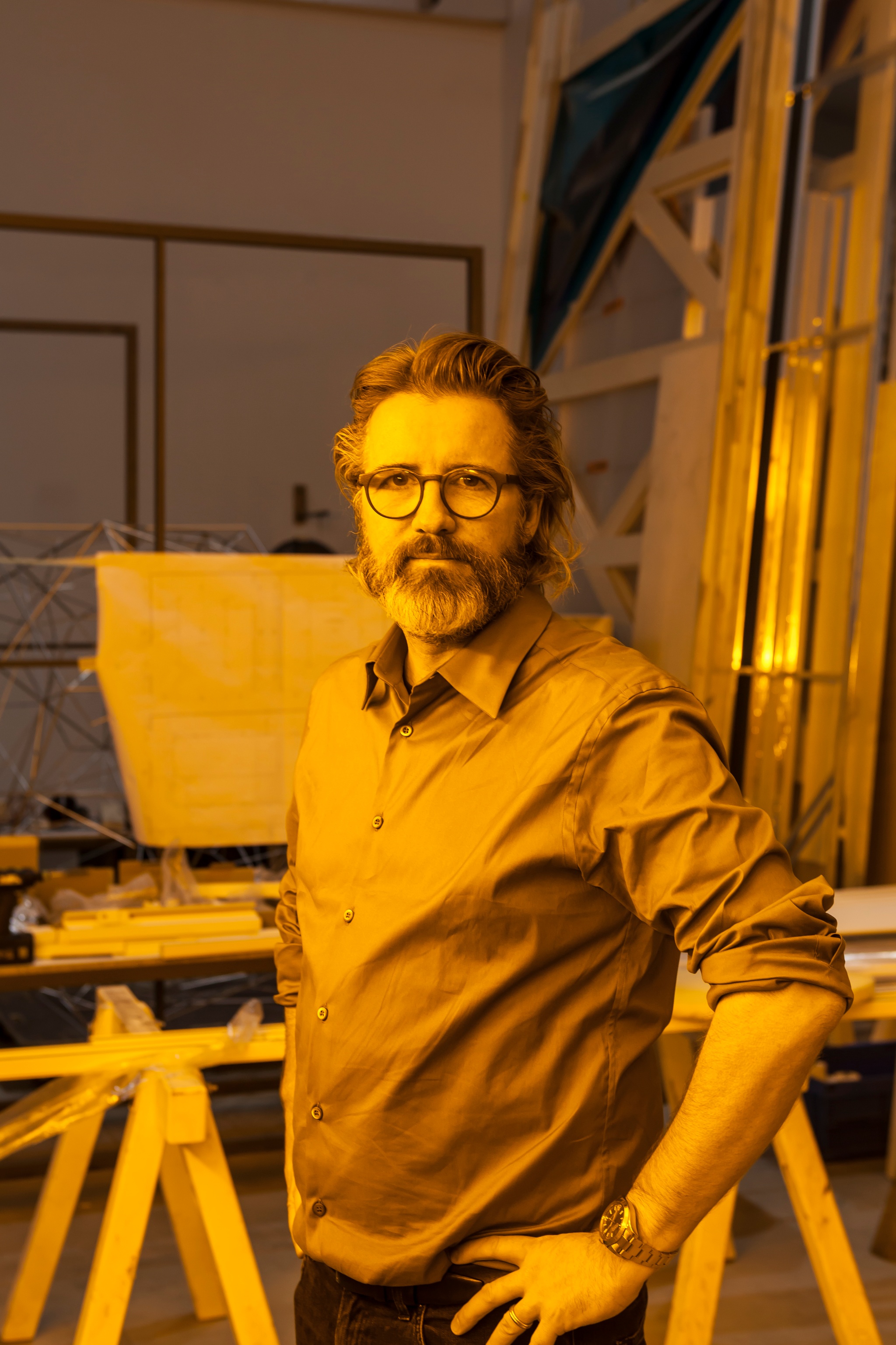 A white man in glasses with a medium length gray beard and long hair swiped back stands in a studio space with hands on his hips.