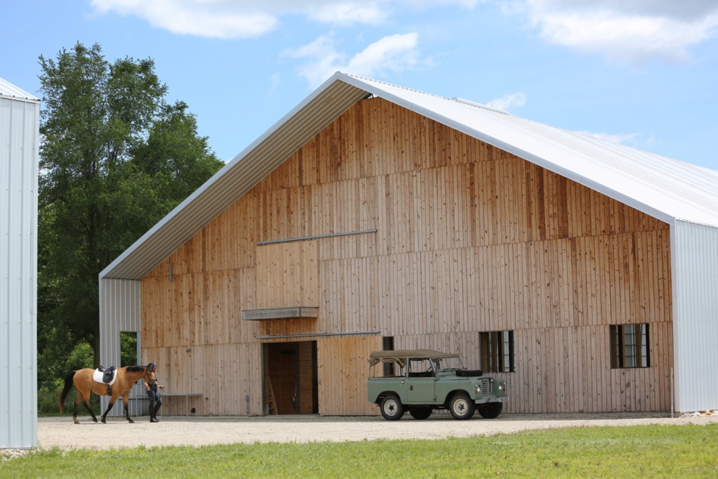 Front of a gabled barn with white roof and siding overhanging the entryway. The front wall is constructed of four rows of vertical wood slats and includes a sliding barn door and two small windows.