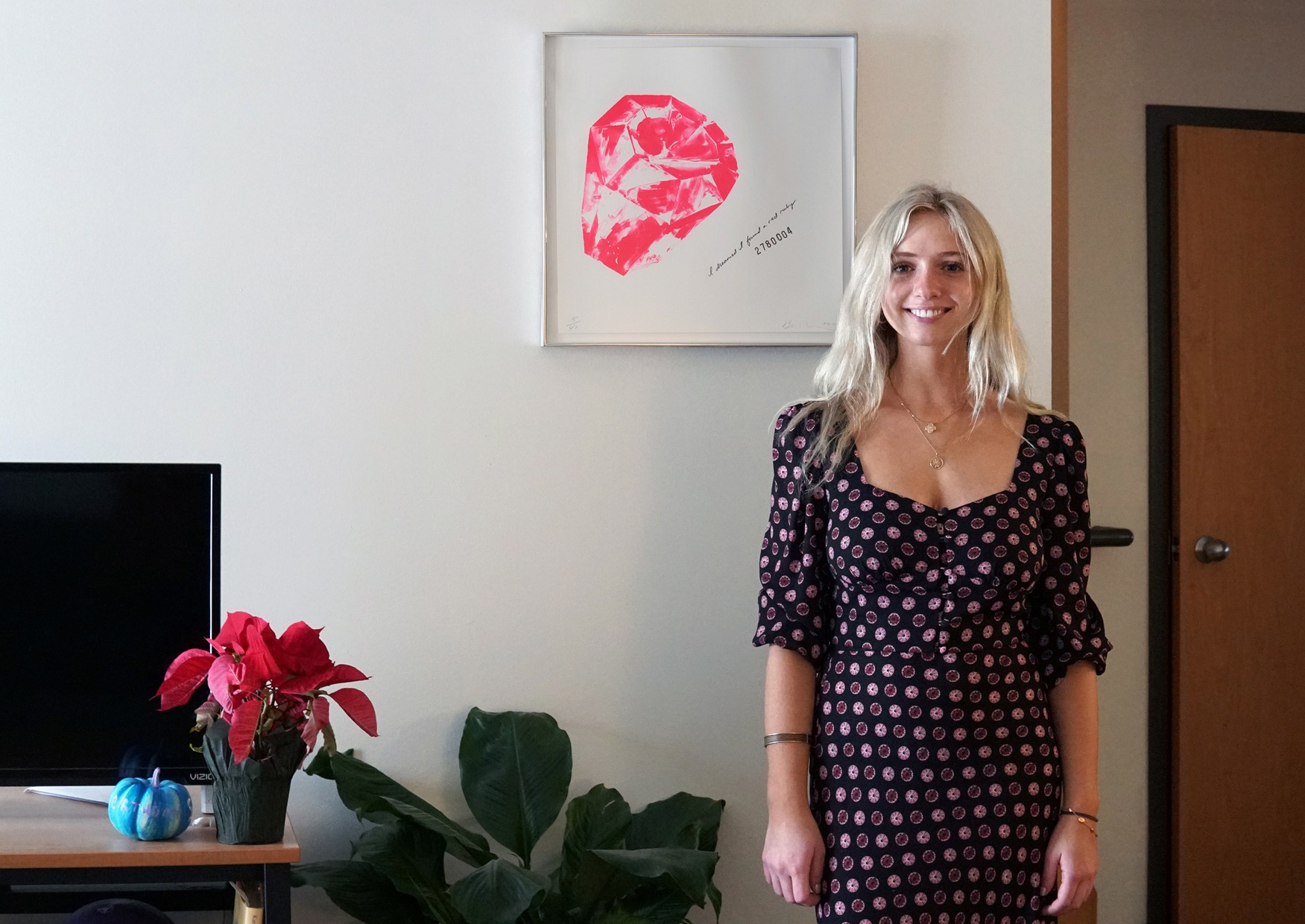 A student stands in her living room next to a framed artwork hung on the wall. The artwork has a white background with a hot pink diamond-like shape in the center left