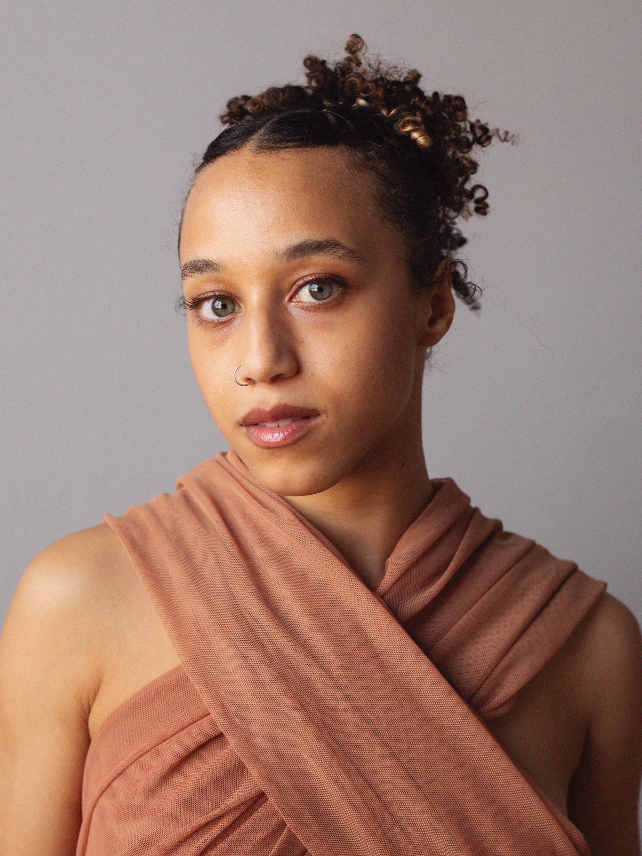 A portrait of Dominican Greene, a biracial, Black, queer woman. She looks directly at us, posing against a neutral background. She has light brown skin and her hair pulled up behind her head. She wears a tan garment that wraps behind her neck, leaving shoulders bare. 