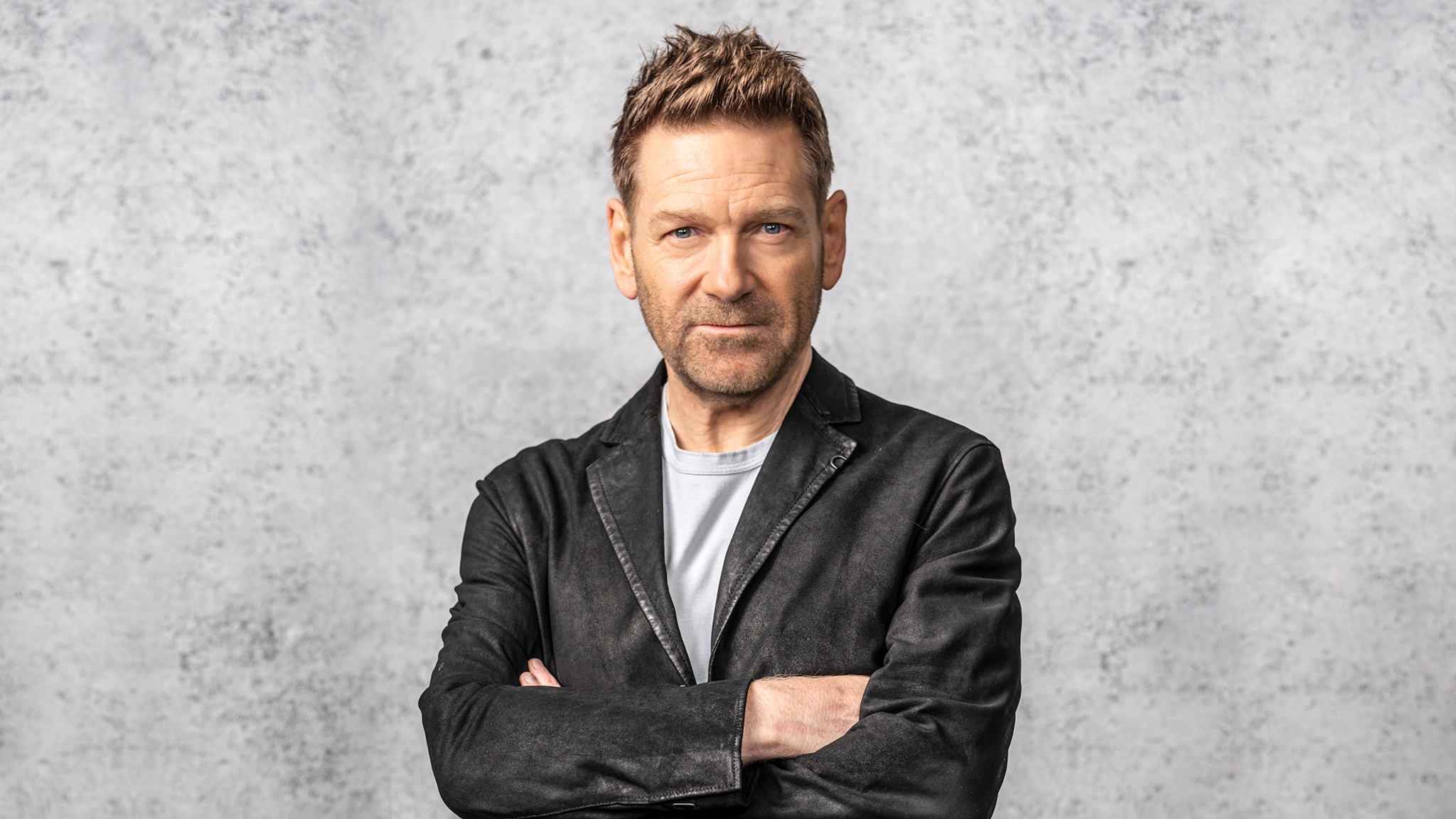 Kenneth Branagh, a white male with brown hair, stands center frame facing forward with his arms crossed. He stands in front of a blurred gray background, in an open black waxed leather blazer and light gray shirt.