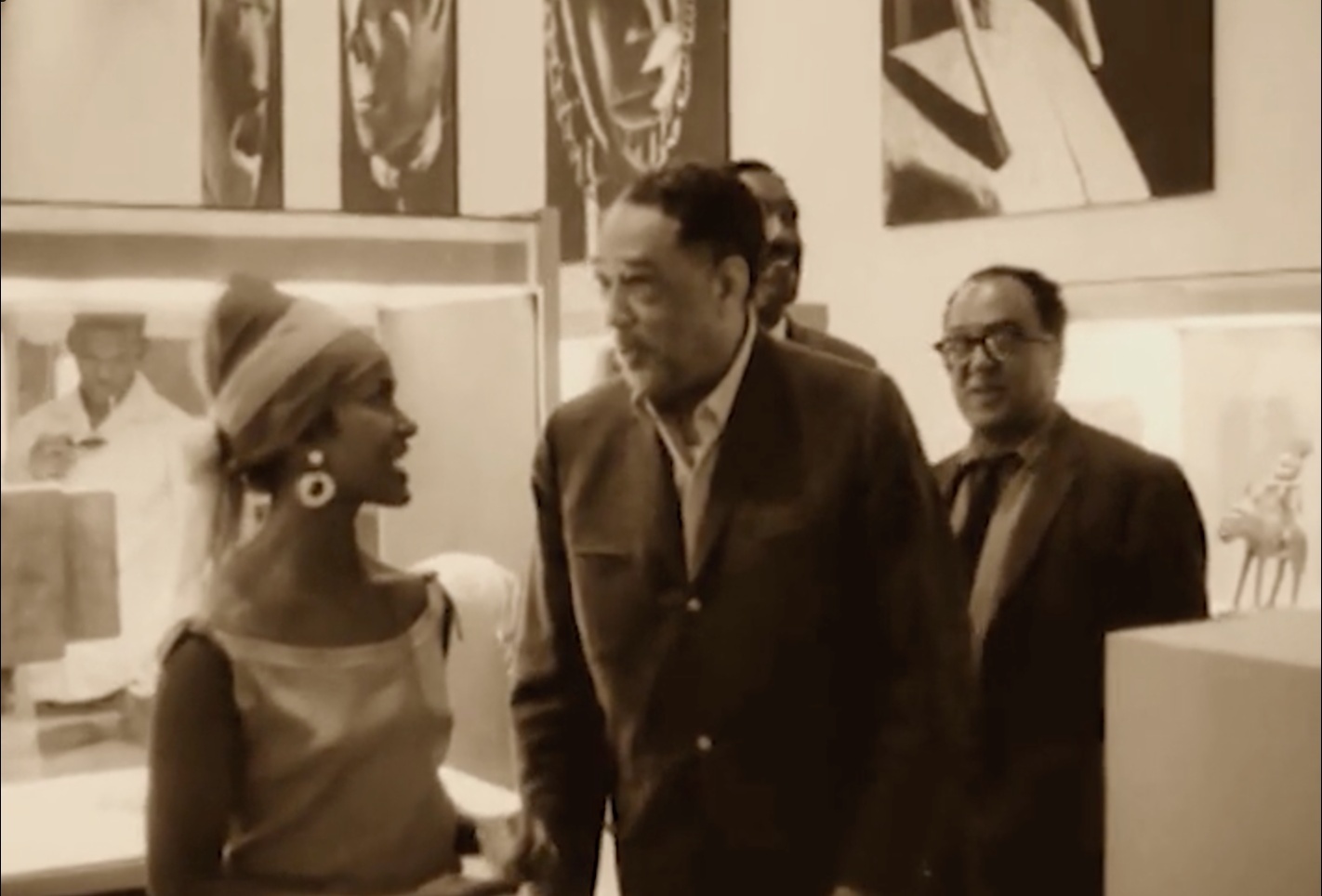 A still from a black-and-white documentary film, showing a woman and three men in a gallery viewing African art