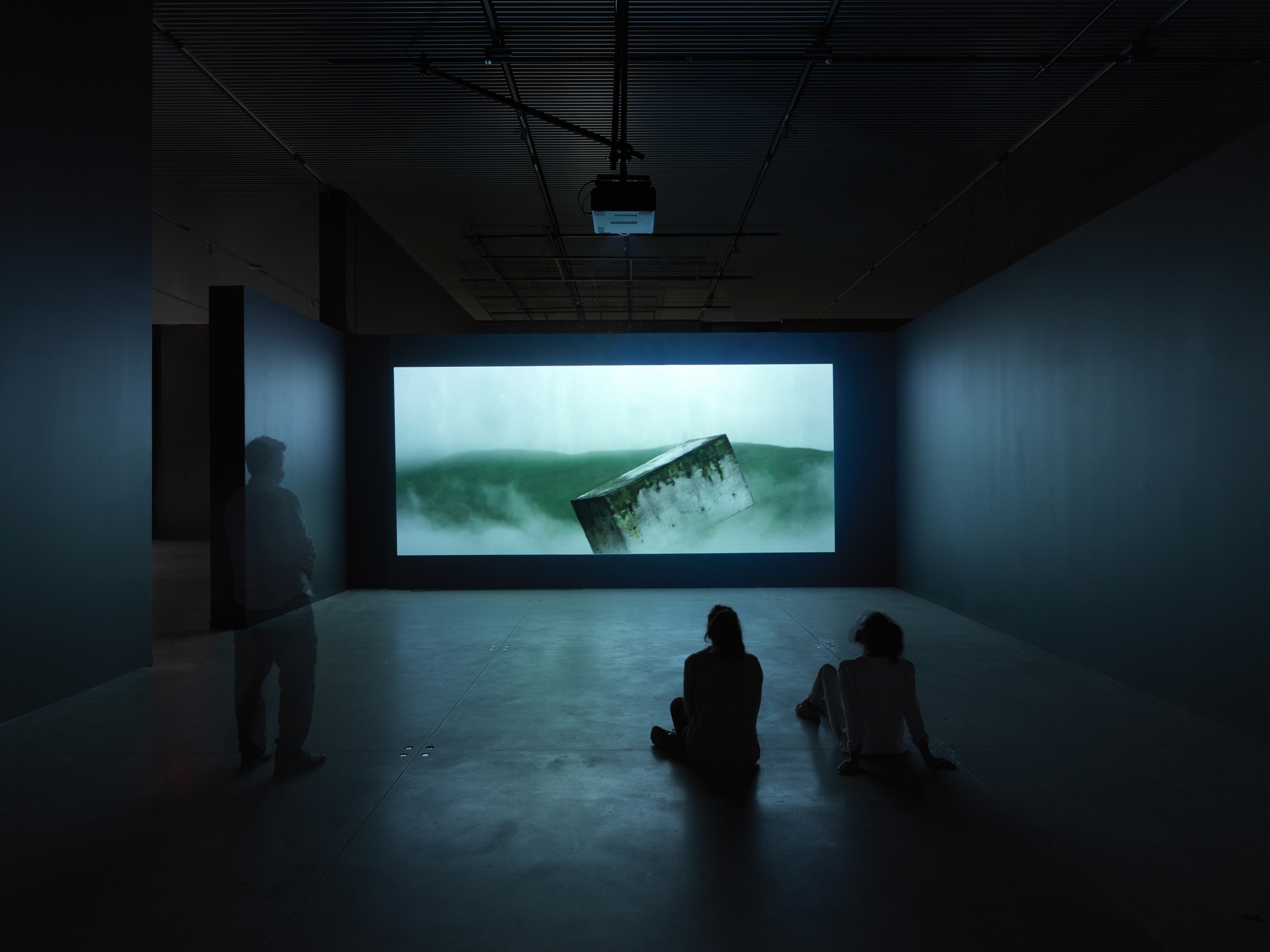 Visitors to The Shed's gallery sit on the floor and stand in front of a film projected on a gallery wall