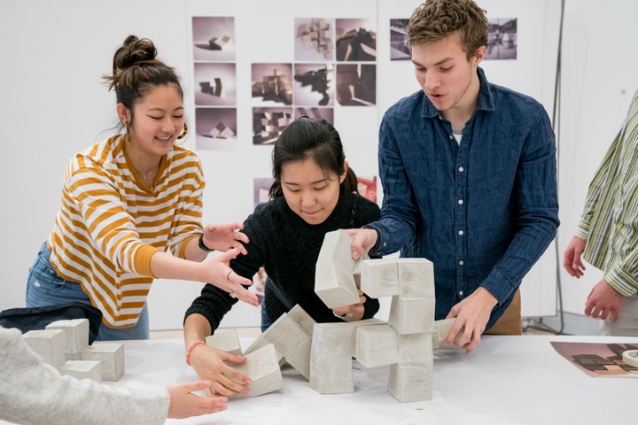 Several students pile modular pieces of concrete into an arrangement on a white table, giggling.
