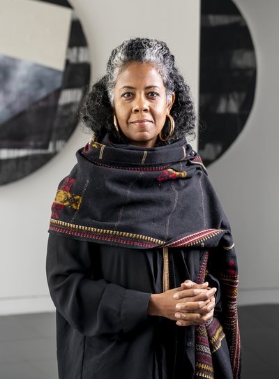 The artist Torkwase Dyson in a black dress with a wide scarf wrapped around her shoulders. Dyson stands in front of artworks hanging on the wall behind her. 