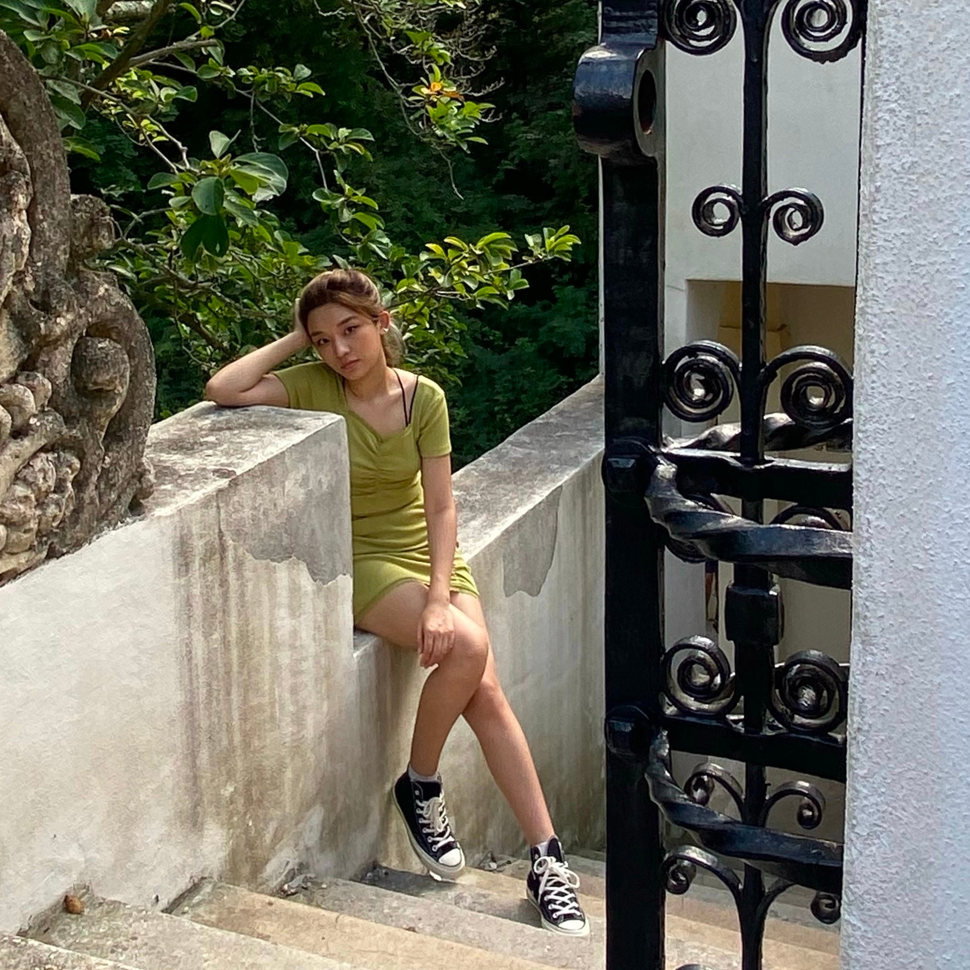 A young Chinese woman wearing a short green dress and black sneakers with white laces sits on a retainer wall that runs along a cement staircase in an outdoor garden setting. 