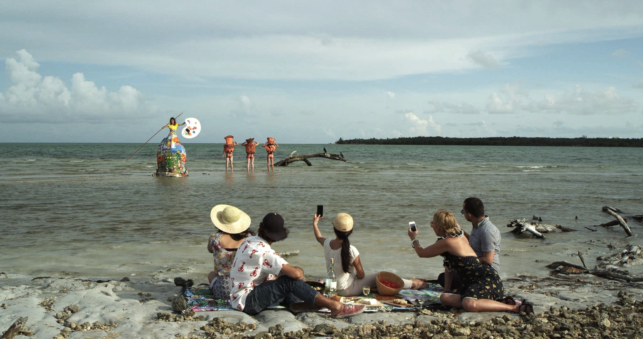 Five adults on a beach hold their phones up towards the water where three people in orange costumes stand next to one person emerging from a colorful structure holding a giant paint palette and stick.