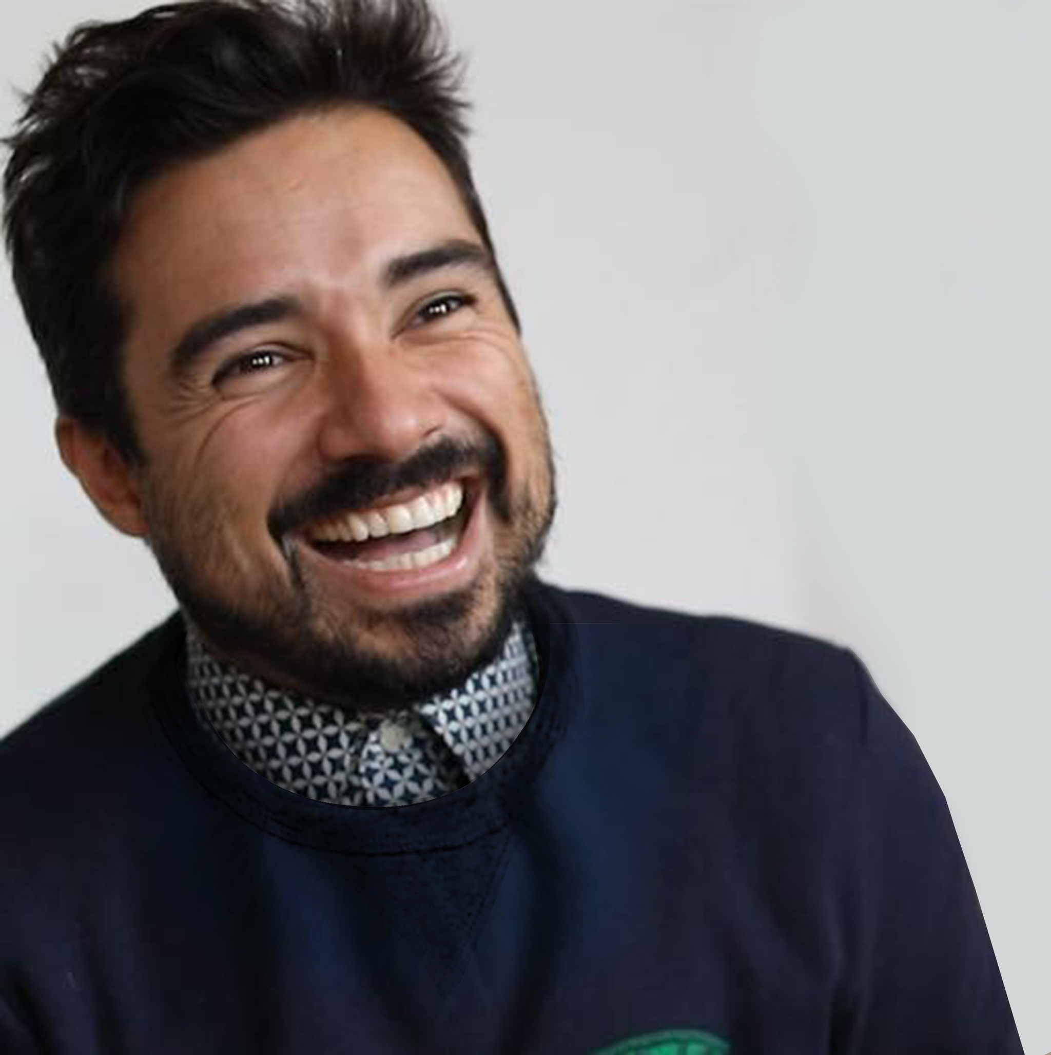 A portrait of Yazmany Arboleda, a Colombian American man who wears a blue sweater with a collared shirt beneath it. He laughs and looks to the side of the image. 