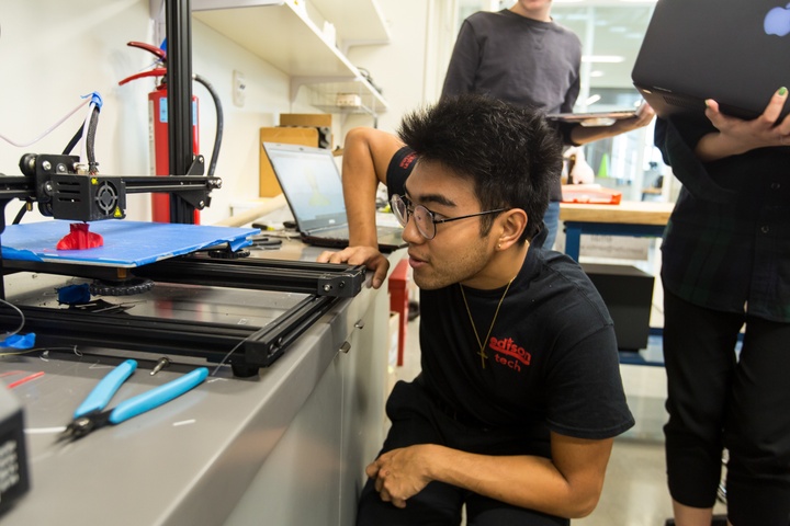 Person watches a 3D printer printing a little model of their head in red.
