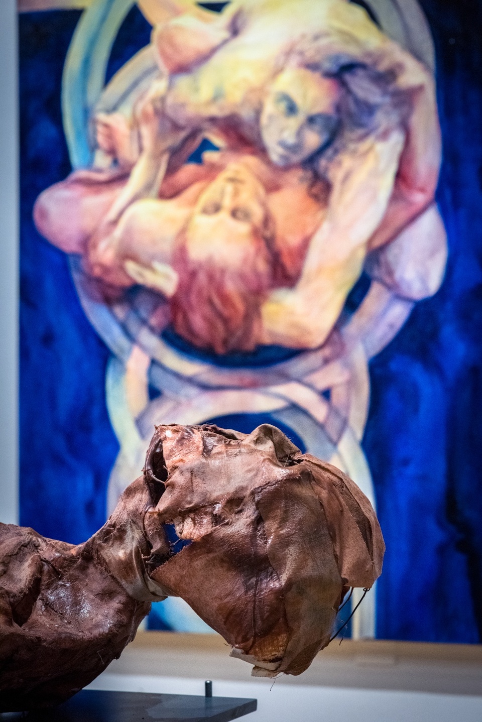 Detail of a bright blue and pinkish painting of two bodies entwined. In front of the painting is a close-up of a leathery-looking mummy head.
