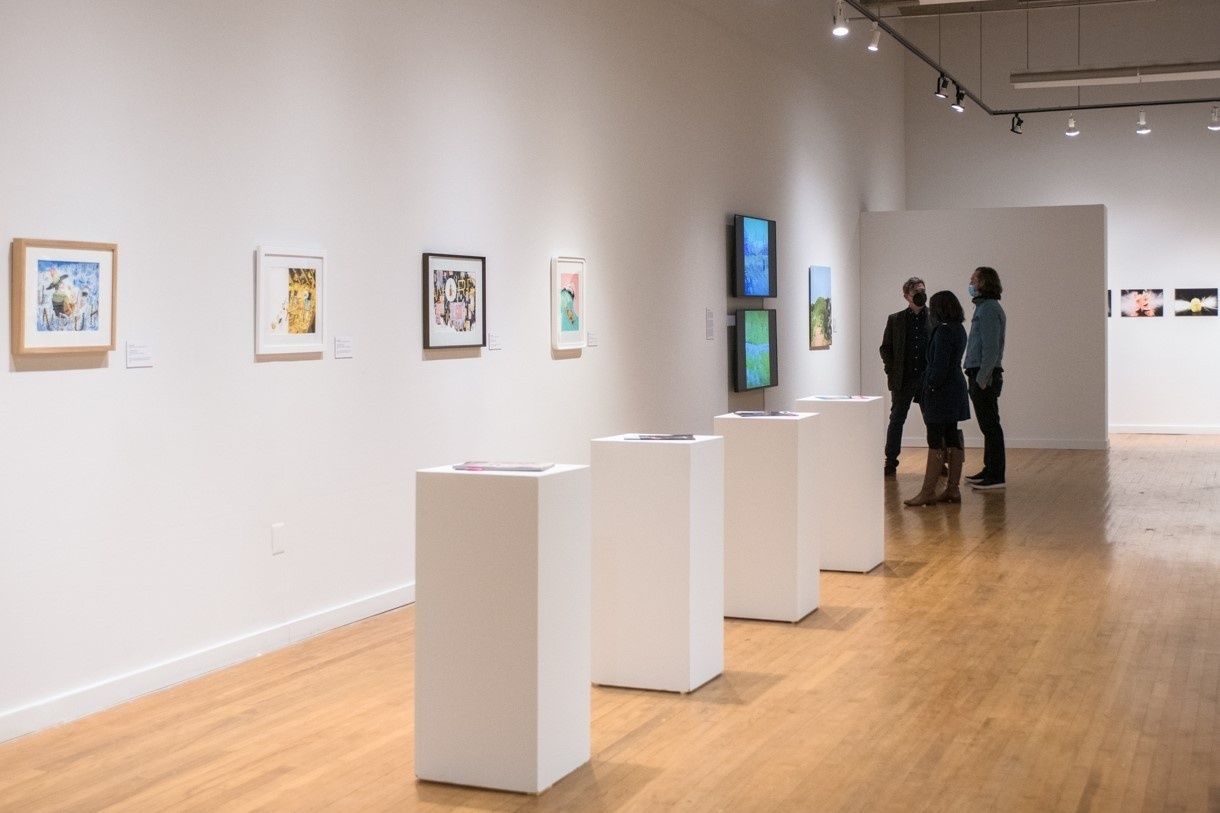 Visitors in a gallery. There are 3 pedestals in the middle with publications and prints in frames on the wall