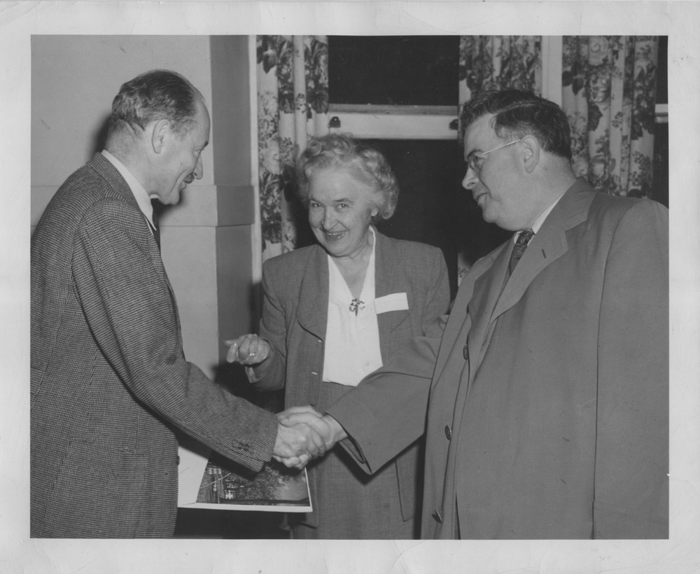 A black and white photograph depicts an older light-skinned woman standing behind two middle-aged light-skinned men in suits shaking hands. The woman looks at the camera while the men look at each other.