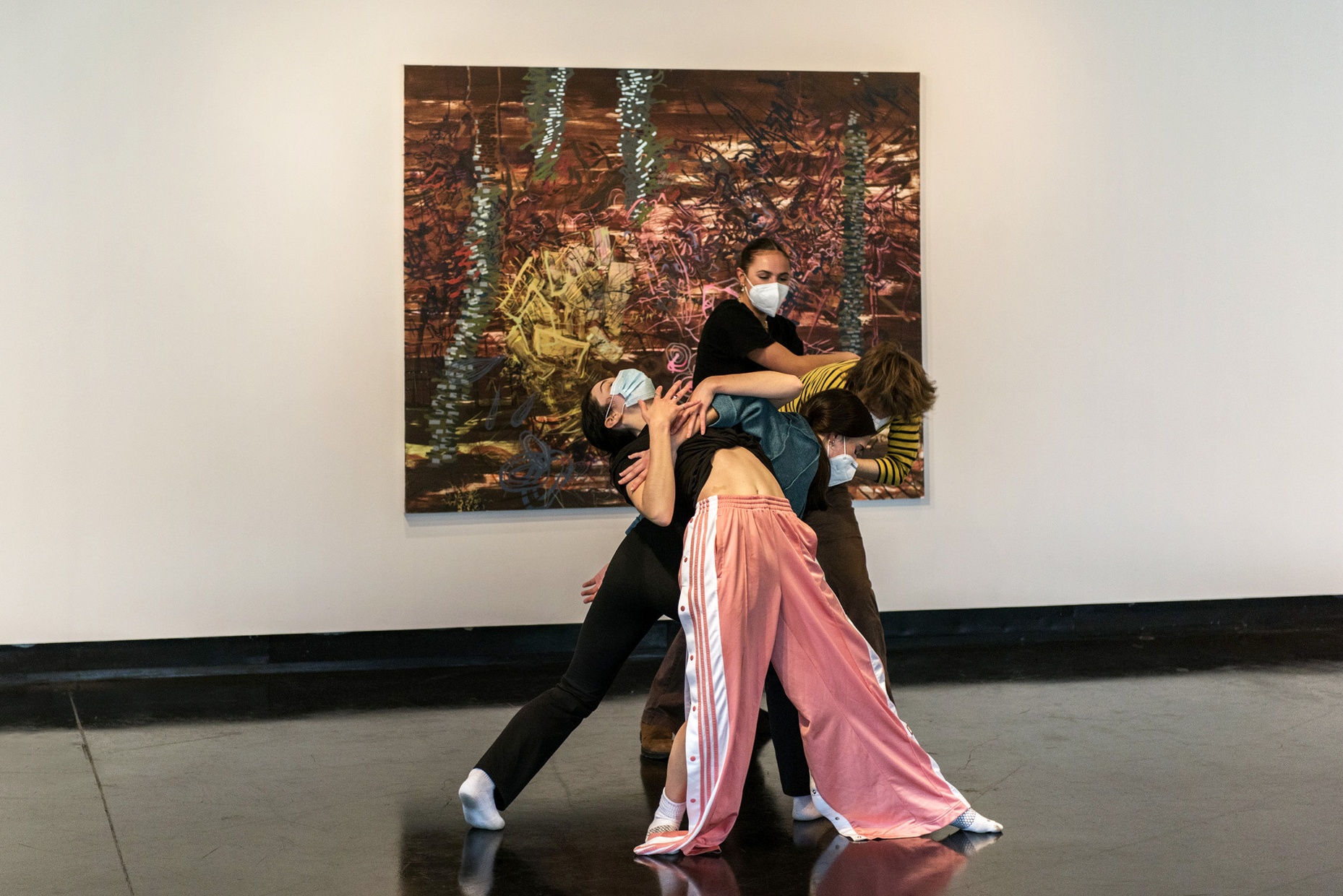 Four masked people dance together in front of an abstract painting hung on the wall behind them.