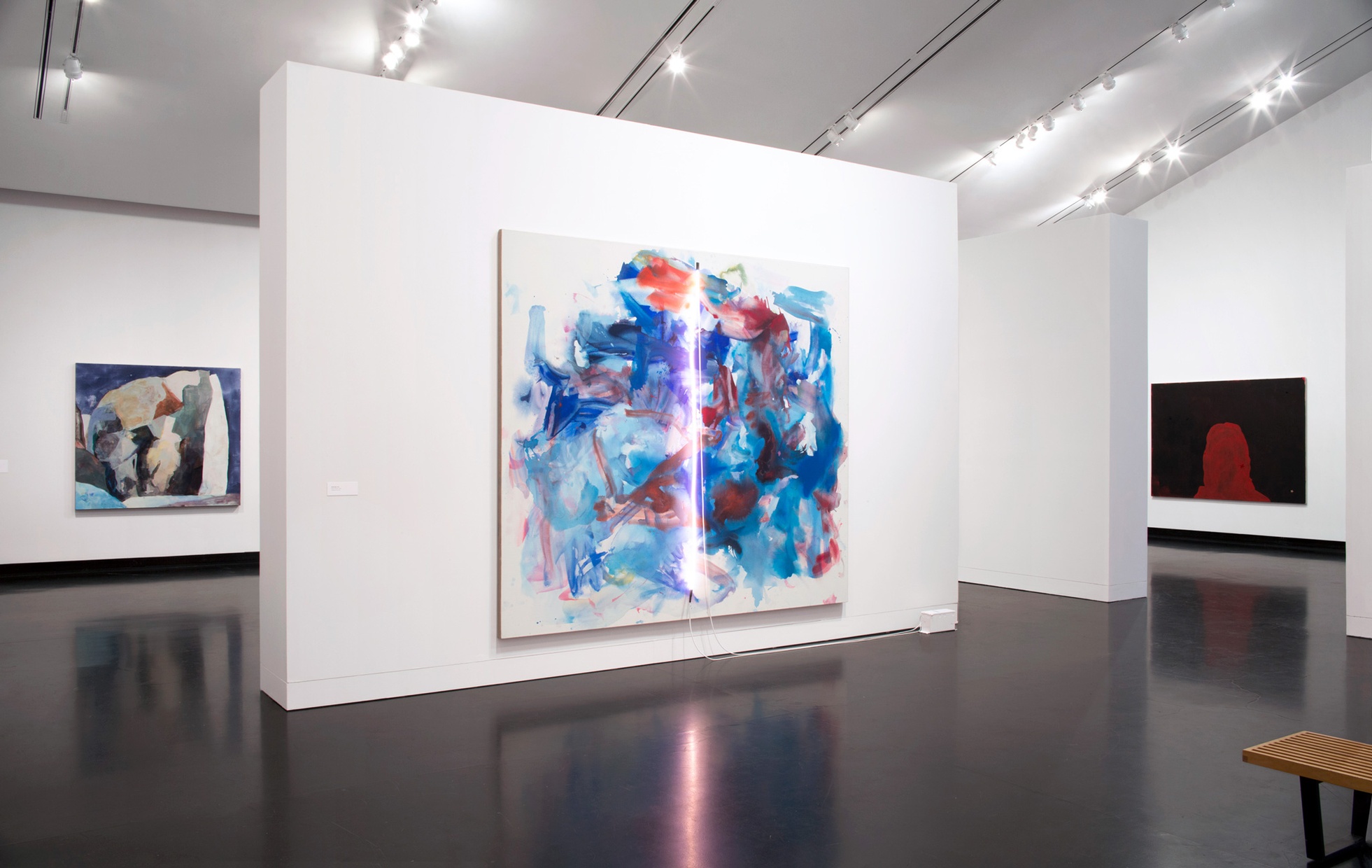 A large, abstract, blue and red painting with a pink, neon tube across the center of the canvas on a white wall with two abstract paintings on walls behind it.