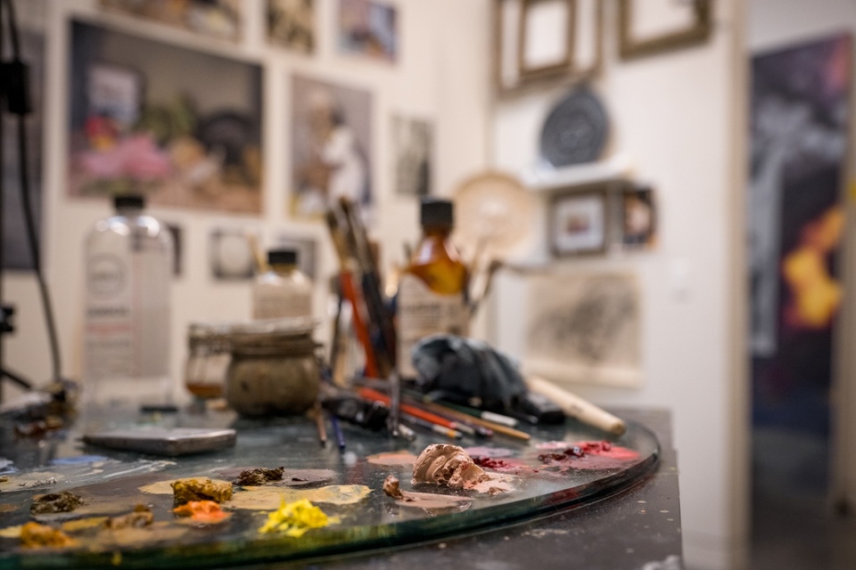 Close up of a paint palette with blobs of brown, yellow, pink, and tan paints. The background is a studio space with an arrangement of paintings in these colors affixed to the wall. 