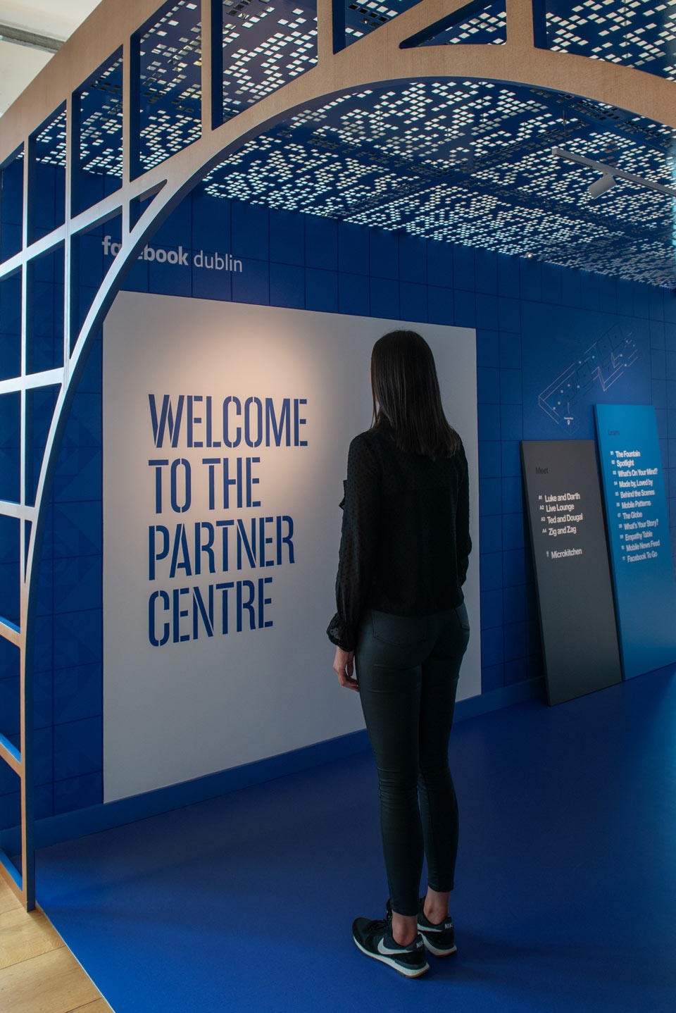 Young woman looking at a sign that says "Welcome to the Partner Center" underneath a logo for "Facebook Dublin", on a blue floor and wall, underneath a grid-like canopy structure