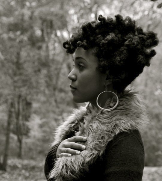 A Black woman standing in profile wearing a sweater with a high and wide furry collar. She stands in a park and in the background are blurry trees.